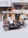 A motorized rickshaw is moving quickly down a street. The blurred background suggests motion and speed. The rickshaw is carrying a person who is bundled up in warm clothing, with decorative cushions on the passenger seats.