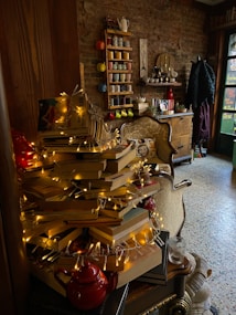 A cozy corner with a stack of books arranged like a Christmas tree, adorned with string lights. There is a teapot at the base of the books. The background features a brick wall with a shelf holding colorful mugs and cups. A vintage-style chair and wooden furniture are also present, creating a warm, inviting atmosphere.