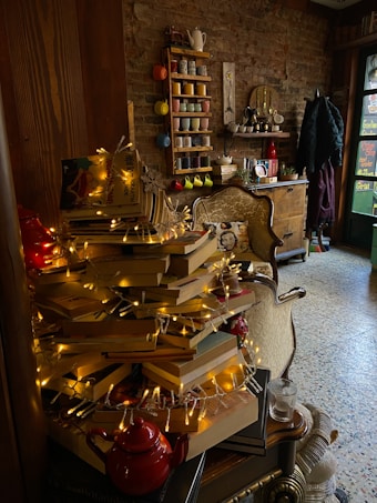 A cozy corner with a stack of books arranged like a Christmas tree, adorned with string lights. There is a teapot at the base of the books. The background features a brick wall with a shelf holding colorful mugs and cups. A vintage-style chair and wooden furniture are also present, creating a warm, inviting atmosphere.