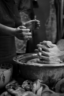 Hands are actively shaping clay on a pottery wheel, demonstrating the art of pottery-making. Two sets of hands are visible, one guiding and the other learning. The scene is intimate and focused, showcasing a transfer of skills.