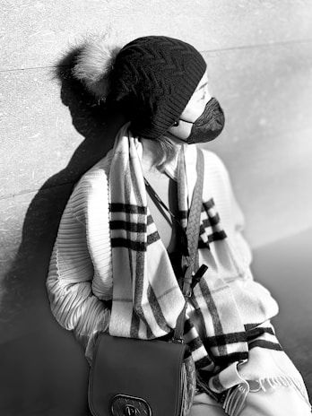 A person wearing a knit hat with a fur pom-pom, a face mask, and a patterned scarf. They are leaning against a textured wall and carrying a leather handbag.