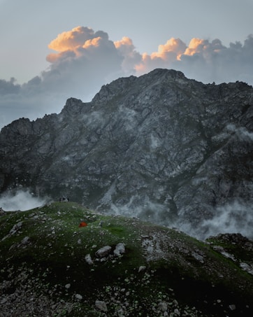 A rugged mountain landscape bathed in soft lighting, with dramatic clouds tinged with orange hues hovering above. Mist rises gently from the ground, lending a mystical atmosphere to the scene. On a grassy hill in the foreground, a small orange tent and a few people are visible, suggesting a remote camping adventure.