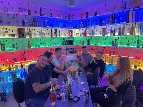 A group of five people are sitting around a table in a bar or lounge. Behind them is a wall filled with bottles of various spirits arranged by color. The lighting creates a vibrant, multicolored backdrop. The table is set with various drinks and snacks, and the people appear to be enjoying a relaxed social gathering.