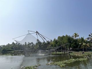 A large fishing net, known as a Chinese fishing net, is suspended over a body of water with lush green trees and palm trees in the background. The sky is clear and blue, and there are patches of aquatic plants floating on the water's surface.
