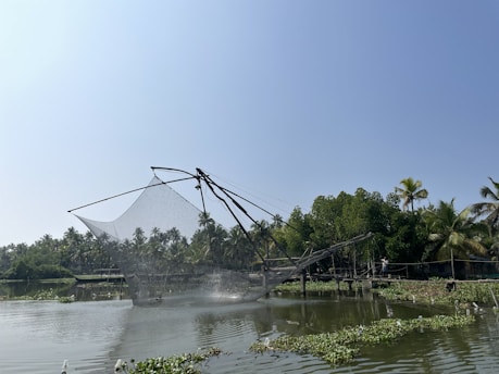 A large fishing net, known as a Chinese fishing net, is suspended over a body of water with lush green trees and palm trees in the background. The sky is clear and blue, and there are patches of aquatic plants floating on the water's surface.