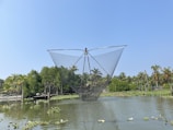 A large fishing net is suspended over a body of water, possibly part of a traditional fishing apparatus. In the background, there are lush green trees and palm trees under a clear blue sky. The water is calm, reflecting parts of the surrounding environment.