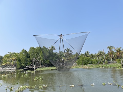 A large fishing net is suspended over a body of water, possibly part of a traditional fishing apparatus. In the background, there are lush green trees and palm trees under a clear blue sky. The water is calm, reflecting parts of the surrounding environment.