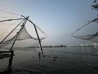 Large fishing nets are suspended over the water using wooden structures at dusk. The horizon is visible in the distance, with lights from a distant shoreline reflecting off the calm water surface.