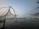 Large fishing nets are suspended over the water using wooden structures at dusk. The horizon is visible in the distance, with lights from a distant shoreline reflecting off the calm water surface.