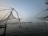 Large fishing nets are suspended over the water using wooden structures at dusk. The horizon is visible in the distance, with lights from a distant shoreline reflecting off the calm water surface.
