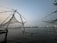 Large fishing nets are suspended over the water using wooden structures at dusk. The horizon is visible in the distance, with lights from a distant shoreline reflecting off the calm water surface.