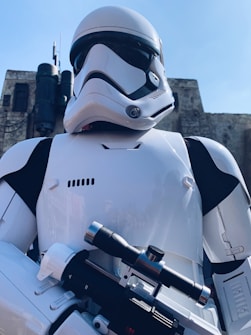 A futuristic character wearing white armored gear stands against a backdrop of industrial buildings. The character has a smooth helmet with a black visor and holds a sleek, futuristic weapon. The setting suggests a science fiction or fantasy theme.