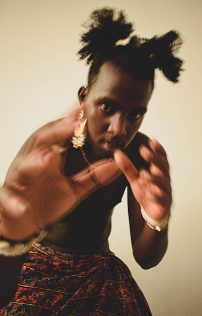 A person with a bold hairstyle featuring two puffs, adorned with gold earrings and bracelets, is captured in a dynamic pose. They appear to be in motion, with hands raised in front, partially obscuring the face. The attire includes a vibrant, patterned wrap or skirt.