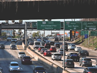 A busy multi-lane highway filled with numerous vehicles in both directions, under several large overhead road signs indicating directions to Hollywood, Ventura, Pasadena, and San Pedro. The scene is set against an overpass and some palm trees and buildings visible in the background.