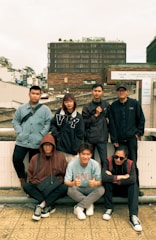 A group of seven young adults is posing outdoors in front of a large modern building with signage that reads 'Dalat Center.' The individuals display a casual style with a mix of sweatshirts, jackets, and glasses. The setting is urban with other buildings visible in the background, and the atmosphere appears relaxed and friendly.