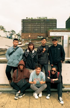 A group of seven young adults is posing outdoors in front of a large modern building with signage that reads 'Dalat Center.' The individuals display a casual style with a mix of sweatshirts, jackets, and glasses. The setting is urban with other buildings visible in the background, and the atmosphere appears relaxed and friendly.