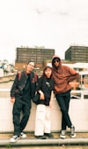 Three individuals are casually standing by a railing, with an urban backdrop featuring modern buildings. They are wearing casual streetwear, including glasses, hoodies, and sneakers. The ambiance feels laid-back and youthful.