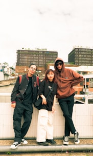Three individuals are casually standing by a railing, with an urban backdrop featuring modern buildings. They are wearing casual streetwear, including glasses, hoodies, and sneakers. The ambiance feels laid-back and youthful.
