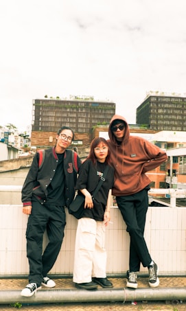 Three individuals are casually standing by a railing, with an urban backdrop featuring modern buildings. They are wearing casual streetwear, including glasses, hoodies, and sneakers. The ambiance feels laid-back and youthful.