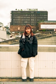 A person stands in a casual pose, wearing a large black sweater with a graphic design and white cargo pants. They are standing against a tiled wall in an urban setting. Behind them, a large building with a modern facade is visible, featuring garden elements on the roof and signs indicating its identity as 'DALAT CENTER'.