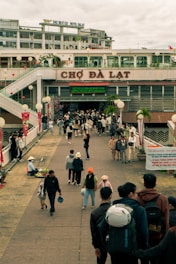 A bustling outdoor market environment with people walking and interacting on a broad walkway. The market entrance is prominent, with 'Chợ Đà Lạt' written above it. There are various shops and signs, and vendors are seen selling goods by the side of the path. The scene includes people of different ages, some carrying bags or helmets, indicative of a busy marketplace setting.