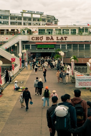 A bustling outdoor market environment with people walking and interacting on a broad walkway. The market entrance is prominent, with 'Chợ Đ&agrave; Lạt' written above it. There are various shops and signs, and vendors are seen selling goods by the side of the path. The scene includes people of different ages, some carrying bags or helmets, indicative of a busy marketplace setting.