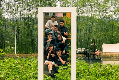 A group of young people pose together within a white frame outdoors, surrounded by lush green vegetation. They are making playful gestures, and some are wearing sunglasses. In the background, there is a bamboo forest and a small structure with a person sitting near a pond.