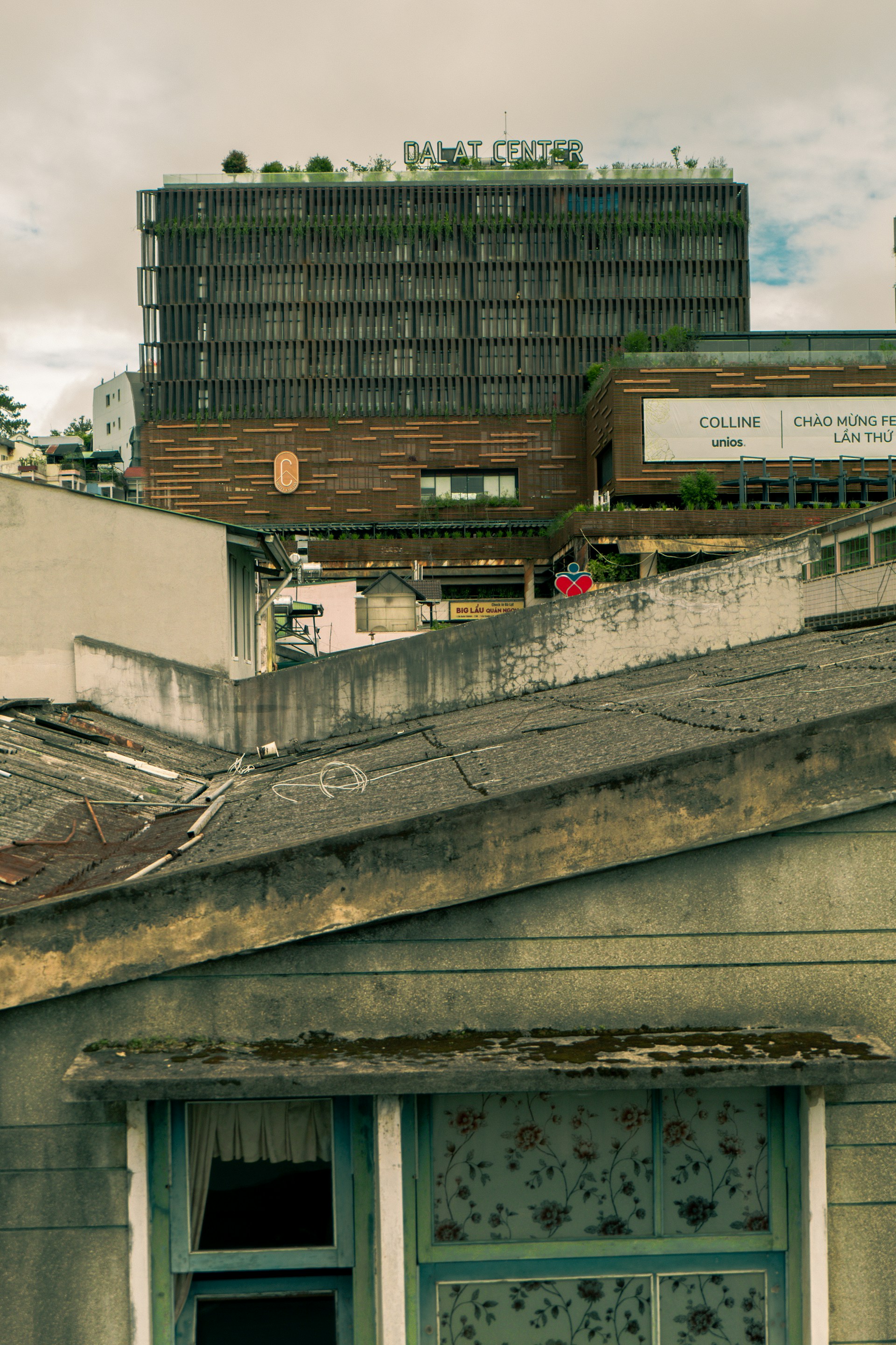 The image features an urban scene with a multi-story building labeled 'Dalat Center' in the background. The facade of the building is modern and textured with greenery on the rooftop. In the foreground, there is a weathered roof with visible wear, including moss and peeling paint. A window with floral-patterned curtains and a partial view of an open window adds to the contrasting mix of the well-kept modern structure and the aged home.