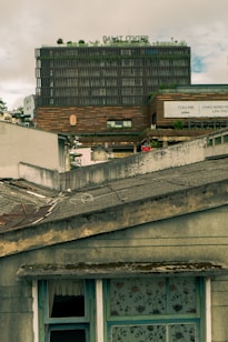 The image features an urban scene with a multi-story building labeled 'Dalat Center' in the background. The facade of the building is modern and textured with greenery on the rooftop. In the foreground, there is a weathered roof with visible wear, including moss and peeling paint. A window with floral-patterned curtains and a partial view of an open window adds to the contrasting mix of the well-kept modern structure and the aged home.