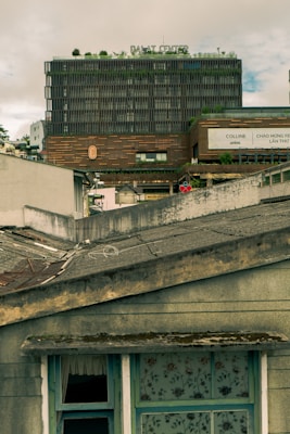 The image features an urban scene with a multi-story building labeled 'Dalat Center' in the background. The facade of the building is modern and textured with greenery on the rooftop. In the foreground, there is a weathered roof with visible wear, including moss and peeling paint. A window with floral-patterned curtains and a partial view of an open window adds to the contrasting mix of the well-kept modern structure and the aged home.