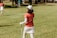 A person in a red shirt and white skirt, with a cap and holding a golf club, stands on a grassy field. Another person in similar attire walks away in the background. The setting appears to be a golf course with trees and golf equipment visible.