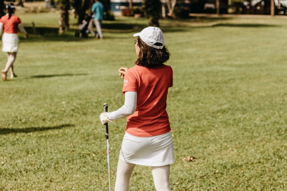 A person in a red shirt and white skirt, with a cap and holding a golf club, stands on a grassy field. Another person in similar attire walks away in the background. The setting appears to be a golf course with trees and golf equipment visible.