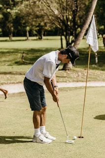 A person in a white polo shirt and dark shorts is playing golf on a green field. They are putting a golf ball towards the hole marked by a flag. The scene is outdoors with lush greenery in the background.