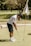 A person in a white polo shirt and dark shorts is playing golf on a green field. They are putting a golf ball towards the hole marked by a flag. The scene is outdoors with lush greenery in the background.