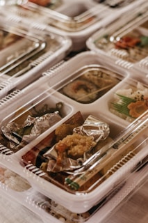 Plastic containers filled with assorted food items, including rice, vegetables, and breaded fried pieces wrapped in foil. The arrangement appears to be a collection of neatly organized meals ready for distribution or consumption.