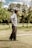 An older man is standing on a golf course with a putter in hand. He is wearing a wide-brimmed hat, a light long-sleeve shirt, and dark pants. The background features trees and a grassy area, suggesting a serene outdoor setting.