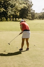 A person is playing golf on a sunny day, wearing a red shirt, white skirt, and a cap. The lush green course is surrounded by trees with a bright, clear sky above.