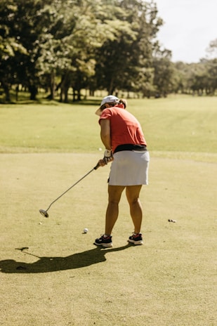 A person is playing golf on a sunny day, wearing a red shirt, white skirt, and a cap. The lush green course is surrounded by trees with a bright, clear sky above.
