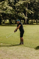 A person is standing on a grassy field, positioned in a golf stance while holding a golf club. They are wearing a blue cap, a black shirt, shorts, and patterned socks. In the background, there are tall trees and a sunlit atmosphere.
