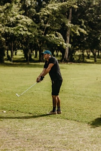 A person is standing on a grassy field, positioned in a golf stance while holding a golf club. They are wearing a blue cap, a black shirt, shorts, and patterned socks. In the background, there are tall trees and a sunlit atmosphere.