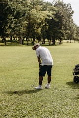 A person is playing golf on a lush green golf course with a golf club in hand. They are in a bent posture, focusing on making a shot. A golf cart is visible nearby, and the background consists of dense trees under a clear sky.
