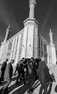 A black and white photograph showing a group of people carrying a coffin draped in fabric, walking toward a large, ornate building with tall minarets. The building's architecture features intricate patterns and arched windows.