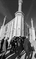 A black and white photograph showing a group of people carrying a coffin draped in fabric, walking toward a large, ornate building with tall minarets. The building's architecture features intricate patterns and arched windows.