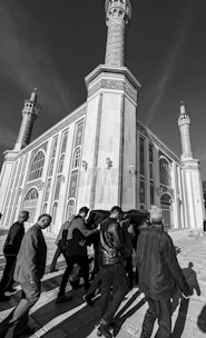 A black and white photograph showing a group of people carrying a coffin draped in fabric, walking toward a large, ornate building with tall minarets. The building's architecture features intricate patterns and arched windows.