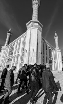 A black and white photograph showing a group of people carrying a coffin draped in fabric, walking toward a large, ornate building with tall minarets. The building's architecture features intricate patterns and arched windows.