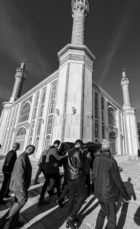A black and white photograph showing a group of people carrying a coffin draped in fabric, walking toward a large, ornate building with tall minarets. The building's architecture features intricate patterns and arched windows.