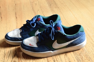 A pair of sneakers placed on a wooden floor with a mixture of blue, green, and white colors. The shoes feature a distinctive swoosh logo on the side, and the laces are tied neatly.