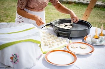 A person is preparing food outdoors on a table covered with a white cloth. Several uncooked dough rounds are placed on a baking sheet, while some are being cooked on a round electric griddle. There are empty orange-rimmed plates, paintbrushes, and cups on the table.