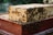 A wooden beehive with numerous bees swarming over it. The hive appears to be well-populated and is constructed from light-colored wood with a reddish base.