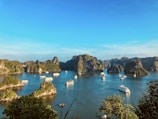 A scenic view of a bay with numerous limestone islands covered in lush greenery, surrounded by calm blue waters. Several boats and cruise ships are scattered across the water, indicating a popular tourist destination. The clear sky and bright sunlight enhance the vibrancy of the landscape.
